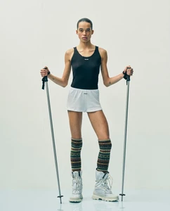 Model holding ski poles wearing a black tank top, white shorts, and patterned knee-high socks against a white backdrop.