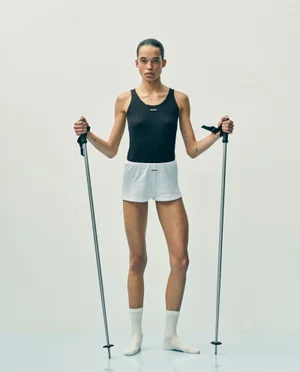 Model Lara Menezes wearing a black tank top and white shorts while holding ski poles against an off-white studio background.