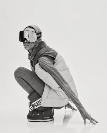 Model Lara Menezes crouching in oversized ski goggles, a puffer vest, ribbed knitwear, and snow boots against a white backdrop.