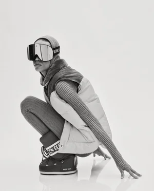 Model Lara Menezes crouching in oversized ski goggles, a puffer vest, ribbed knitwear, and snow boots against a white backdrop.