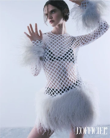Fashion editorial shot of a woman wearing a mesh dress with white fur accents against a clean studio backdrop, showcasing a stylish look.