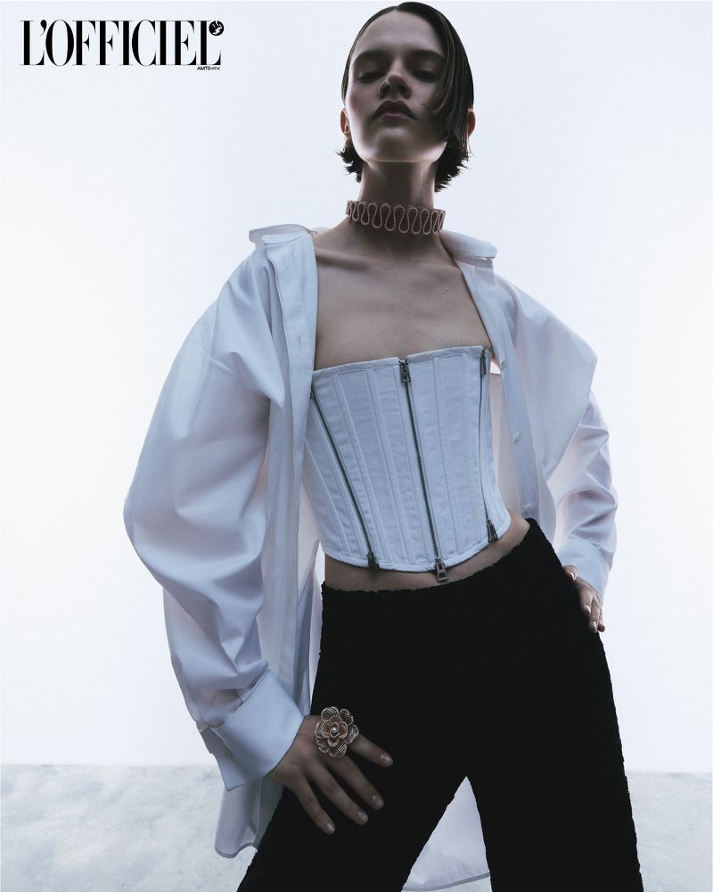 A fashion shot of a young female adult wearing a white corset and a white, oversized shirt with black pants in a studio setting.