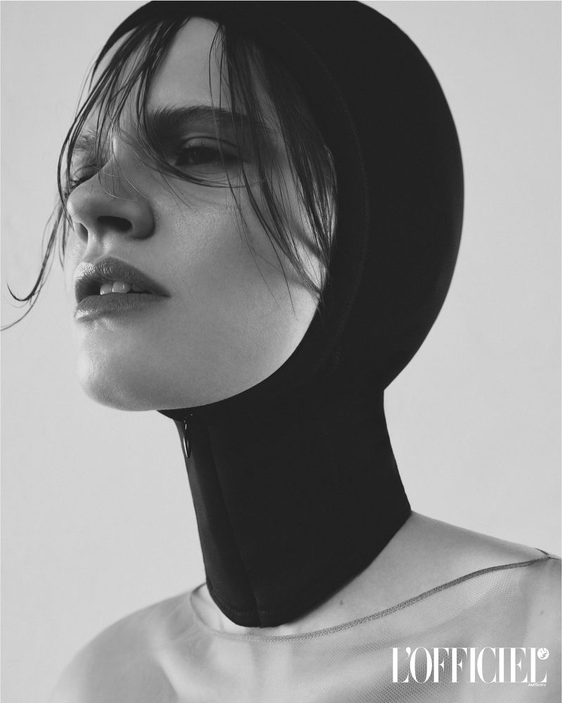 Close-up portrait of a young woman with wet hair wearing a black balaclava and sheer top, set against a neutral gray backdrop.