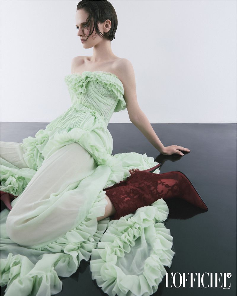 Fashion shot: Woman in ruffled light-green gown, textured brown boots sitting on glossy dark surface against a white background. L'Officiel logo