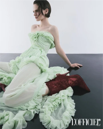 Fashion shot: Woman in ruffled light-green gown, textured brown boots sitting on glossy dark surface against a white background. L'Officiel logo