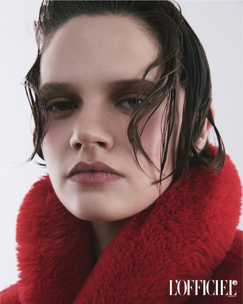 Close-up fashion shot of a model with wet dark hair and red faux fur coat, set against a clean white backdrop with magazine logo.