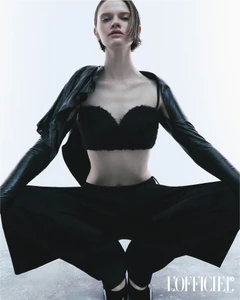 Model Lila Pankova crouching in a black bralette, wide-leg trousers, and leather shrug against a white studio backdrop.