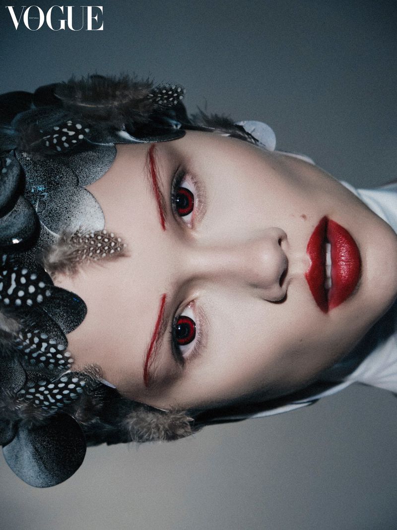 Fashion portrait of a woman with red lips, brows, and eye accents wearing a feather headpiece in a high fashion editorial style.