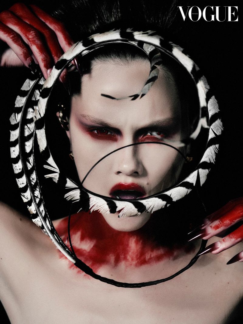 Editorial fashion shot of a model with open mouth, feather boa, red paint, and dramatic makeup against a black background.