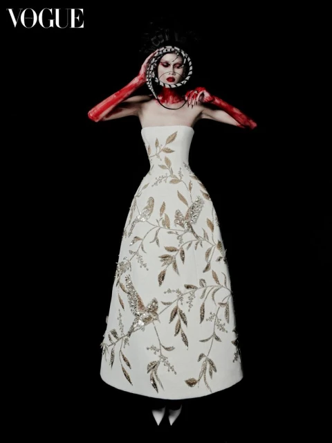 A model wearing a white strapless dress with an elaborate headpiece is center frame against a dark backdrop in a fashion editorial.