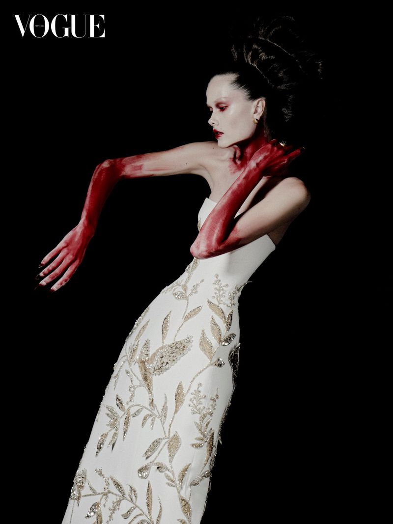 Fashion editorial shot of a woman in a gown, posing against a black background with red paint covering her arms and neck.