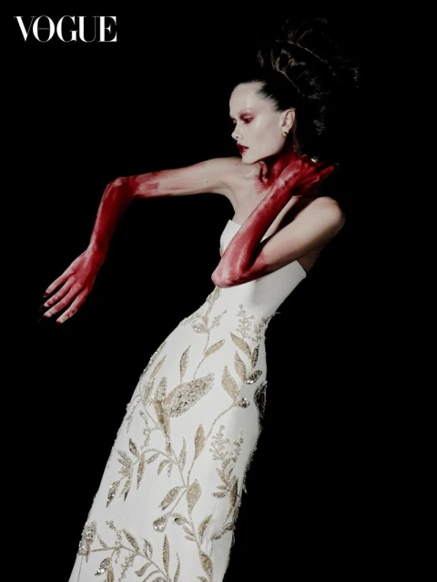 Fashion editorial shot of a woman in a gown, posing against a black background with red paint covering her arms and neck.