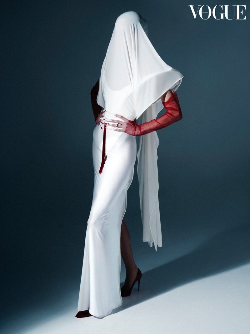 Fashion shot of a woman in a white dress and red gloves posing with a white drape covering her head against a grey background.