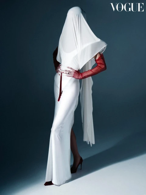Fashion shot of a woman in a white dress and red gloves posing with a white drape covering her head against a grey background.