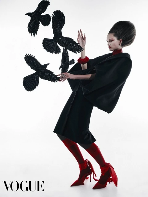 A woman in a black outfit poses with black bird props against a bright background in a fashion editorial. Vogue logo visible.