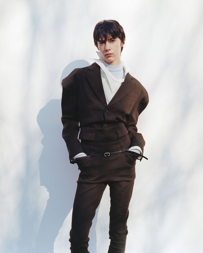 Adult fashion model poses in a dark brown suit with an oversized blazer and matching trousers in front of a white wall with shadows.