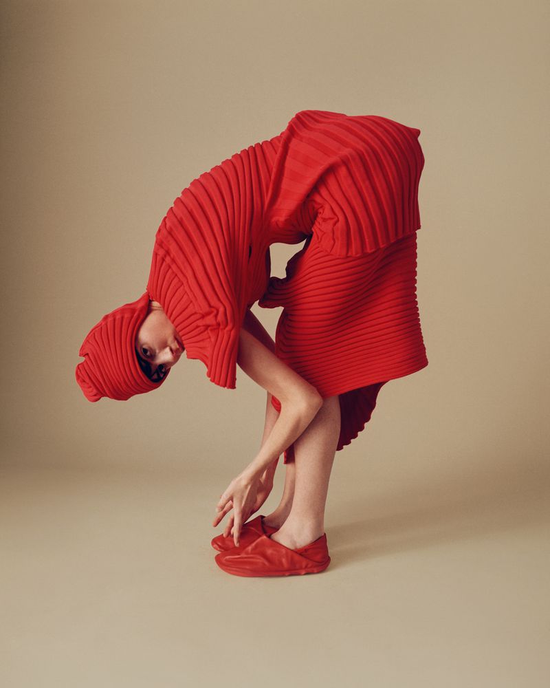 Woman in red pleated dress and matching shoes bending over in a studio with a light beige backdrop, showcasing fashion and design.