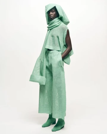 Fashion model with dark skin in mint green hooded outfit and matching scarf, top, pants, and ankle boots, in a photo studio.