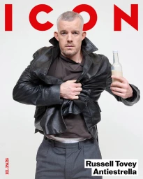 Actor Russell Tovey wearing a black leather jacket over a brown top, holding a glass bottle against a white background.