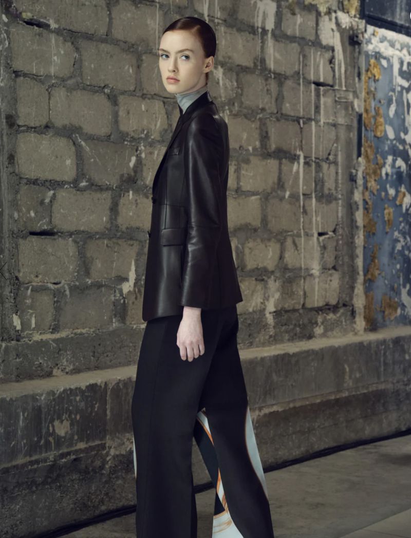 A young fair-skinned woman in a black leather jacket and wide-legged pants poses against a textured concrete wall indoors.