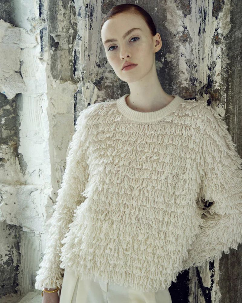 Portrait of a young adult female fashion model in a fringed sweater against a textured wall, looking at the camera with a neutral expression.