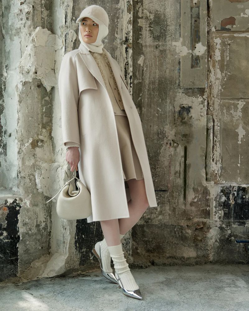 Model in cream coat and matching cap holding handbag against a weathered concrete and stone wall