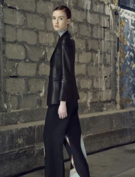 Model in black leather blazer and patterned trousers standing against a textured concrete block wall.