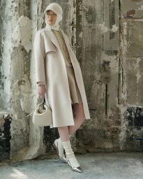 Model in cream coat and matching cap holding handbag against a weathered concrete and stone wall