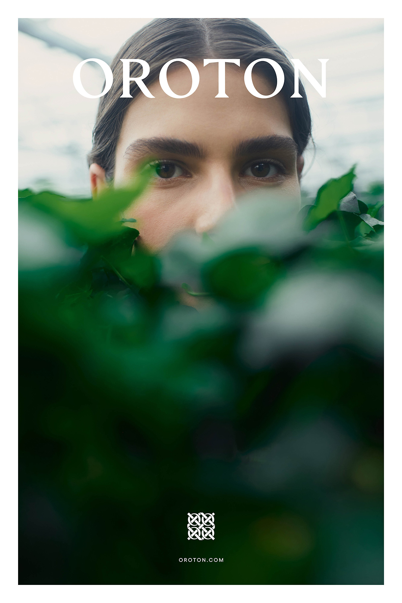 Close-up studio shot features a model partially obscured by lush green foliage, highlighting fashion brand OROTON against a light backdrop.