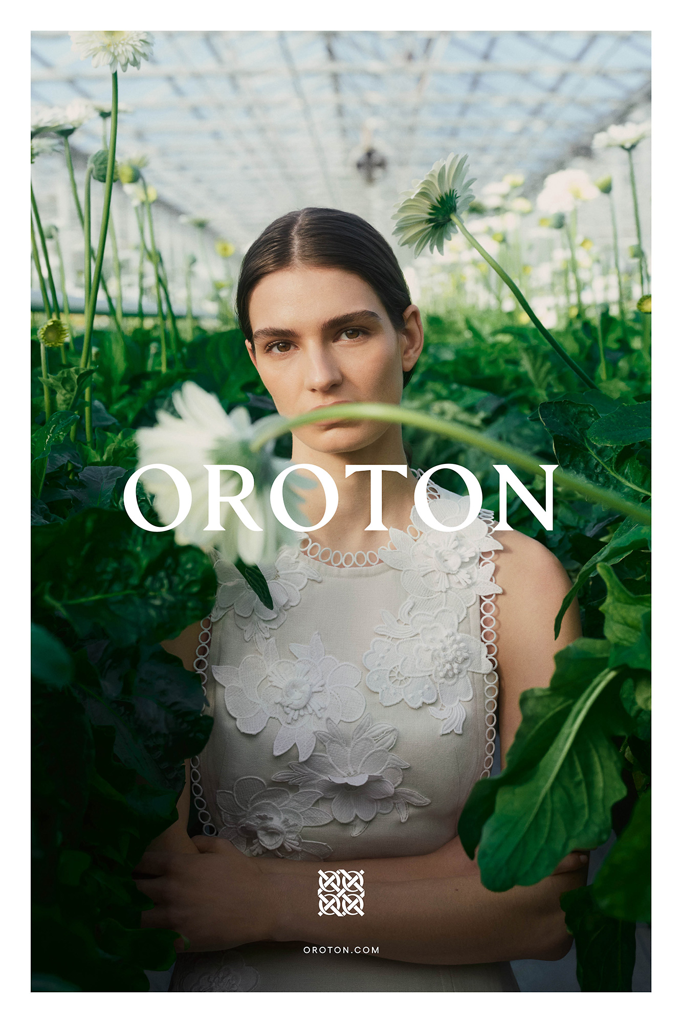 Woman with flowers in fashion advertisement for Oroton, wearing a white lace dress, posing indoors in daylight, and looking at the camera.