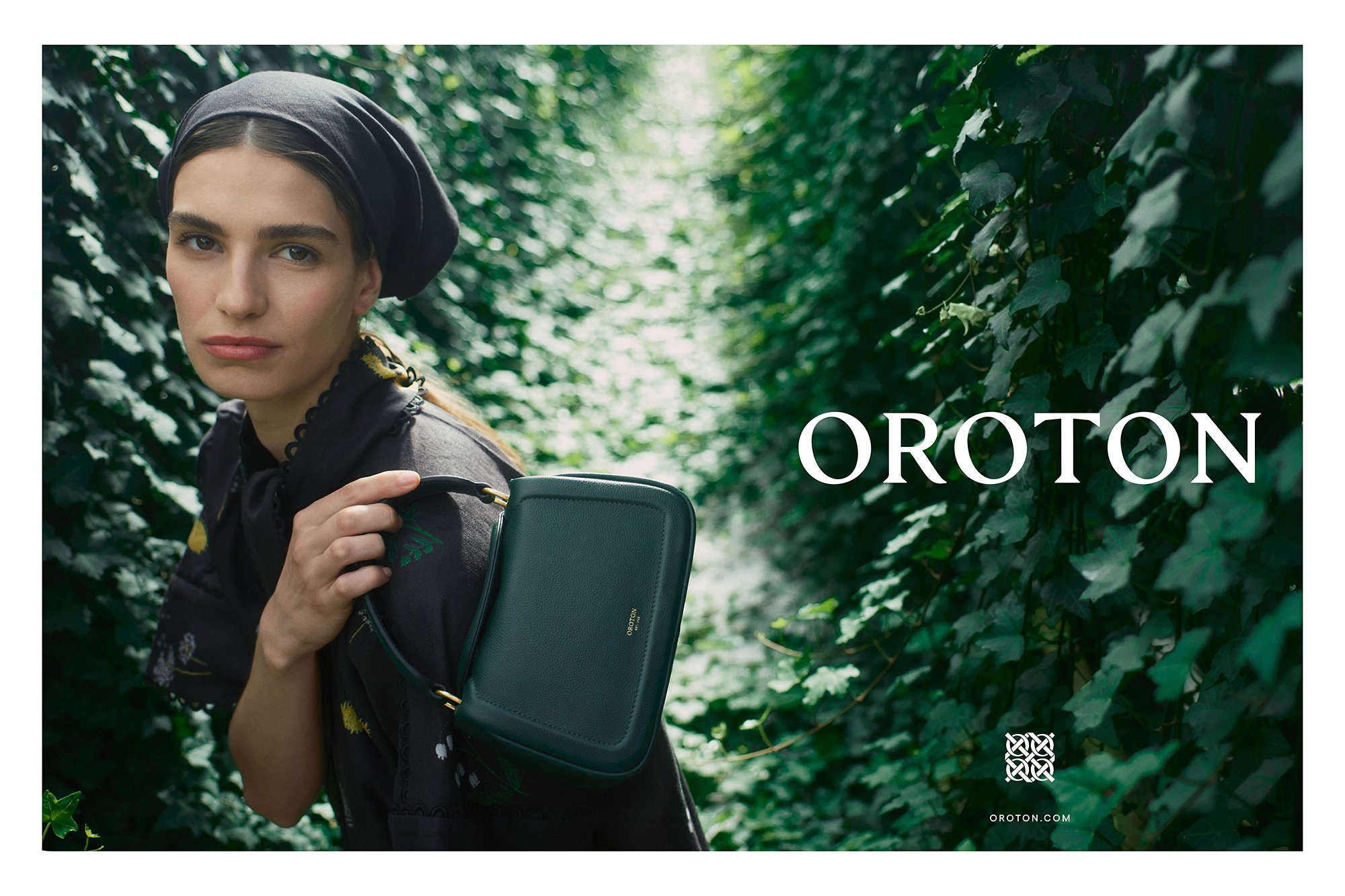 Woman in floral dress, wearing headscarf, holds Oroton handbag in leafy outdoor setting for fashion brand visual marketing.