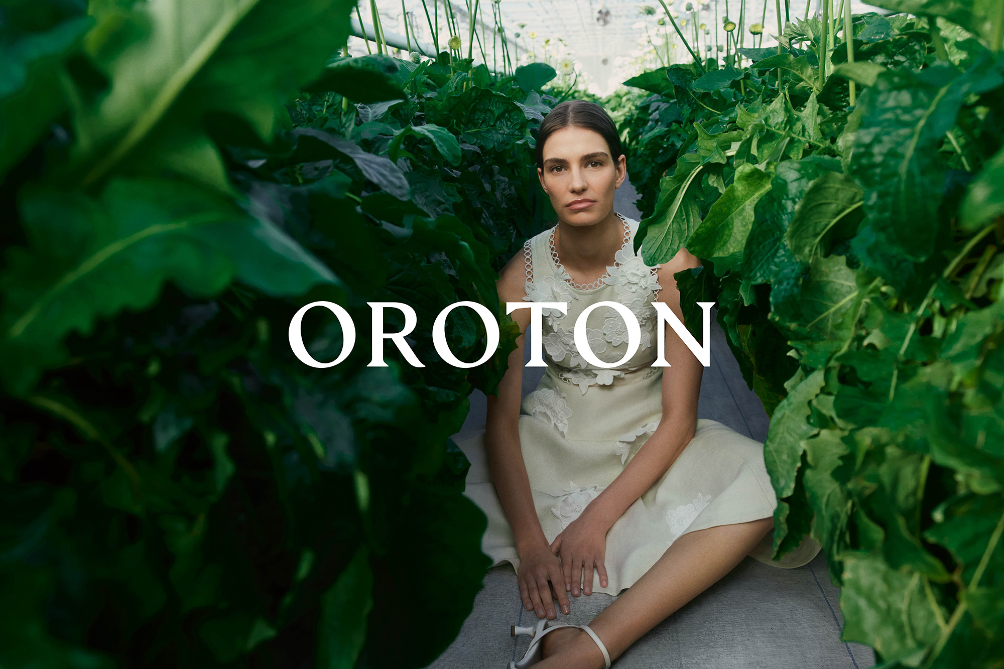Woman in a white floral dress sitting among lush green plants in a greenhouse, fashion editorial advertisement for Oroton brand.