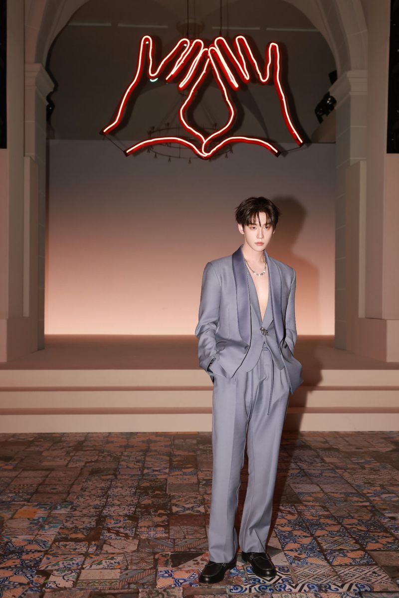 Adult male in a grey suit stands in front of steps and a neon hand art installation in an indoor setting with patterned flooring.
