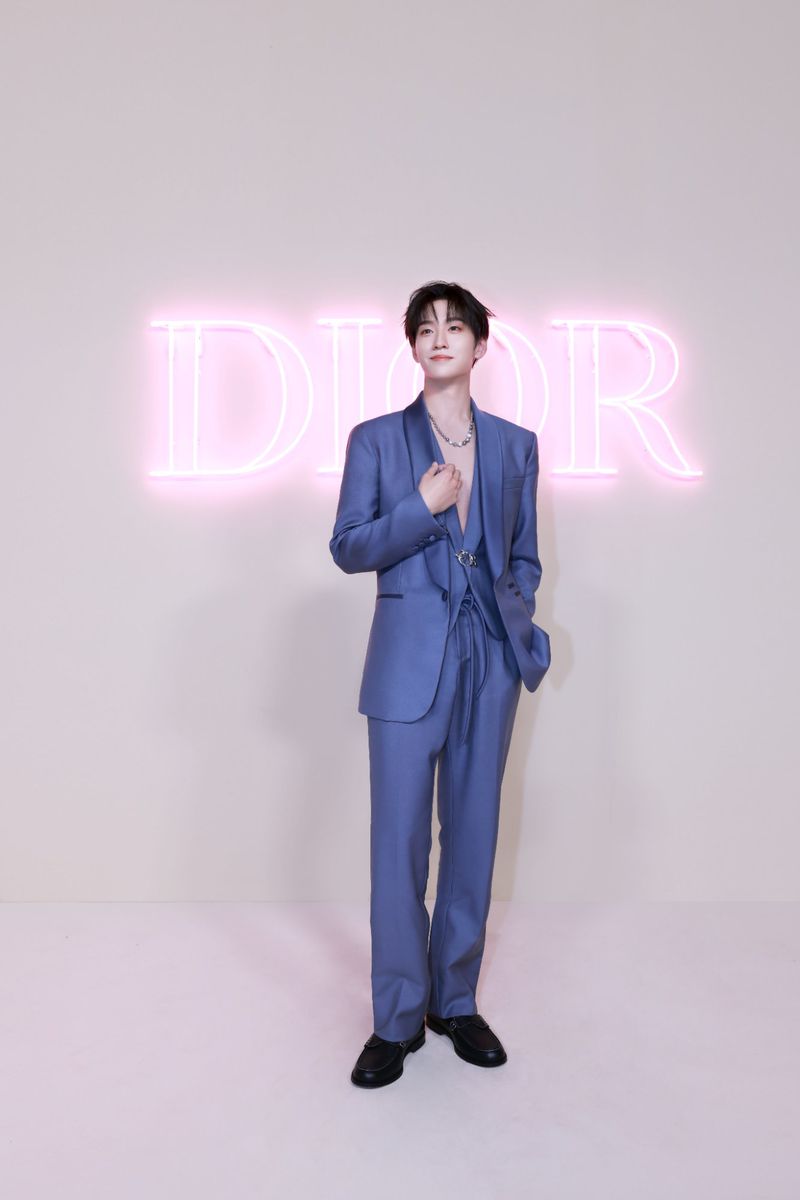 Young adult poses in a blue suit in front of a pink Dior logo, portrait shot, indoor setting, clean background, fashion and entertainment.