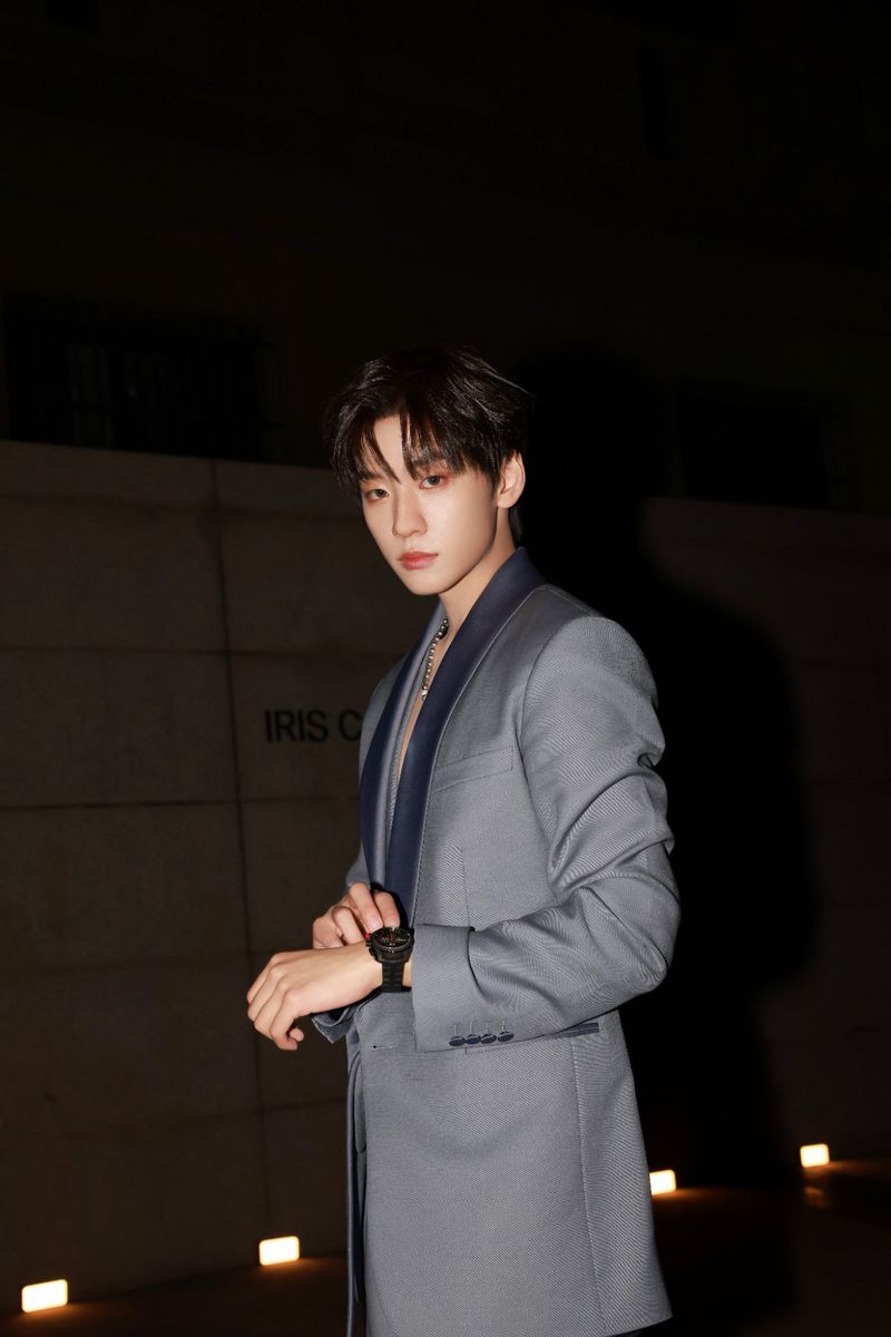 Portrait of an adult male in a suit with dark hair, posing in front of a dimly lit wall with subtle architectural details and ambient lighting.