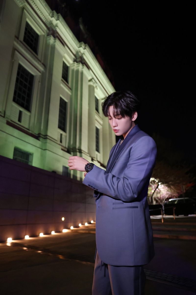 A stylish Asian man in a blue suit stands outside at night, illuminated by ambient light, with an old building as a backdrop.