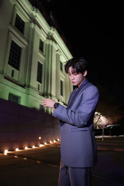 A stylish Asian man in a blue suit stands outside at night, illuminated by ambient light, with an old building as a backdrop.