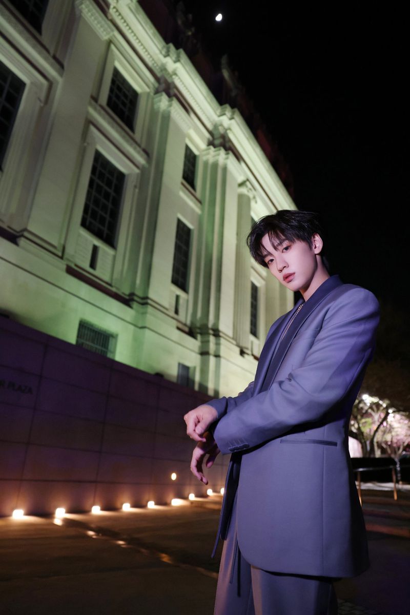 A young adult in a suit stands outside a classic building at night, illuminated by soft lighting, posing with a serious expression.
