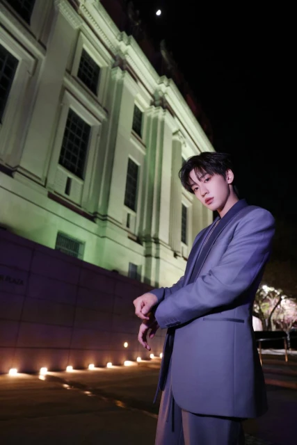 A young adult in a suit stands outside a classic building at night, illuminated by soft lighting, posing with a serious expression.