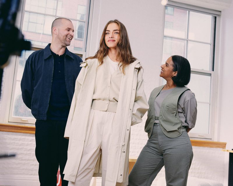 Three people stand indoors in a brightly lit room; an adult female poses as a fitting model as two people look at her.