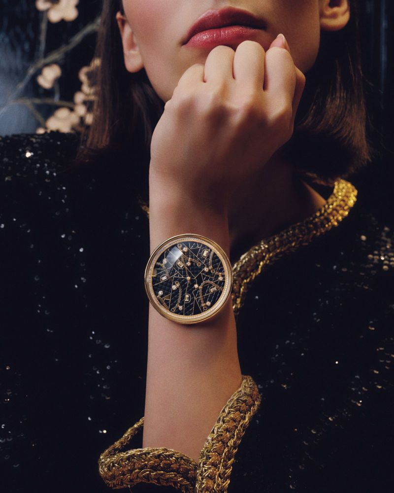 A woman models a vintage gold chain necklace and an ornamental wristwatch against a dark background with floral accents. Beauty shot.