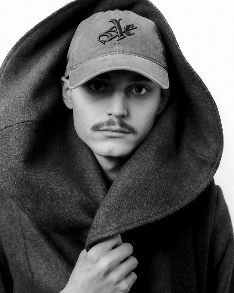 Monochrome portrait of a young adult man in a hooded grey coat and baseball cap against a white backdrop. Striking portraiture.