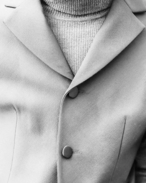 Close-up on a light-colored blazer with two buttons and a ribbed, light gray turtleneck sweater, showing a classic, minimalist fashion style.