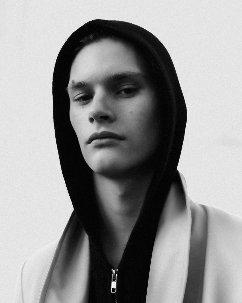Close-up black and white portrait of a young male model wearing a dark hooded sweatshirt against a plain white background.
