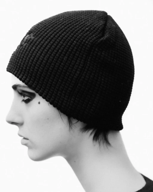 Profile shot of a young adult woman with dark eye makeup and a black knit cap, set against a bright white background, close-up.