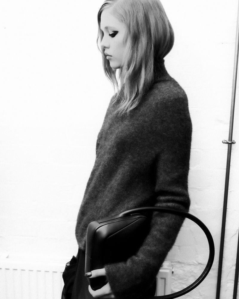 Model in dark fuzzy sweater holding a black structured handbag against a white brick wall