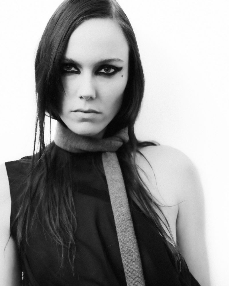 Monochrome portrait of a woman with dark hair, bold eye makeup, and a serious expression wearing a black top and grey scarf against a white background.