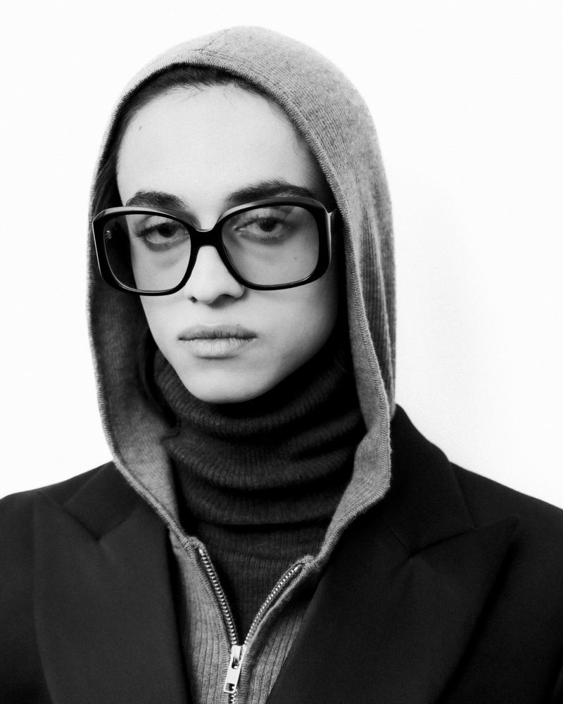 Monochrome portrait of a person wearing a hooded zip-up sweater, turtleneck, blazer, and eyeglasses against a white background.