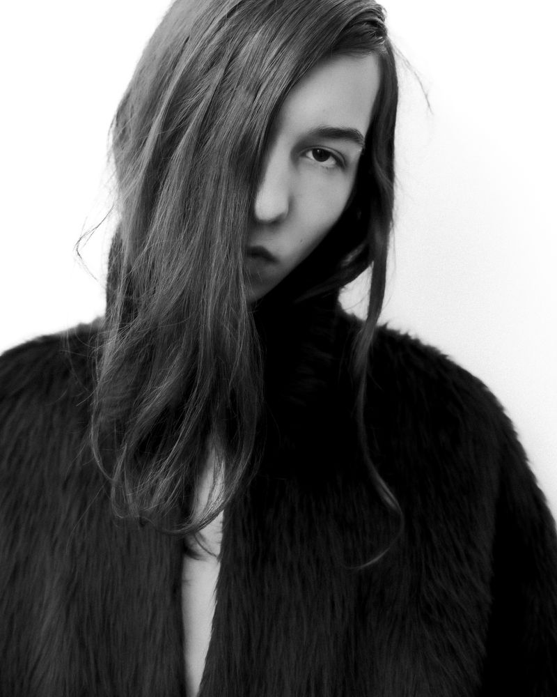 Black and white portrait of a young adult woman with long hair partially obscuring her face, wearing a dark, soft jacket.