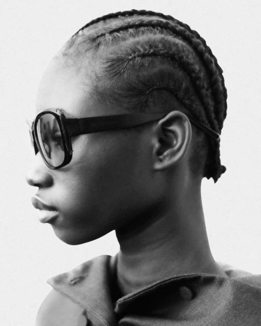 Black and white portrait of African American person with braided hair wearing dark glasses against a neutral background in eye-level shot.